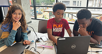 Three middle school students from a previous camp sit at a table withe a laptop computer.