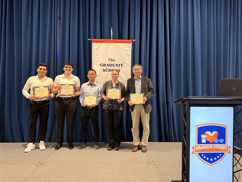 Recipients pose in fron to the Graduate School banner holding their certificates.