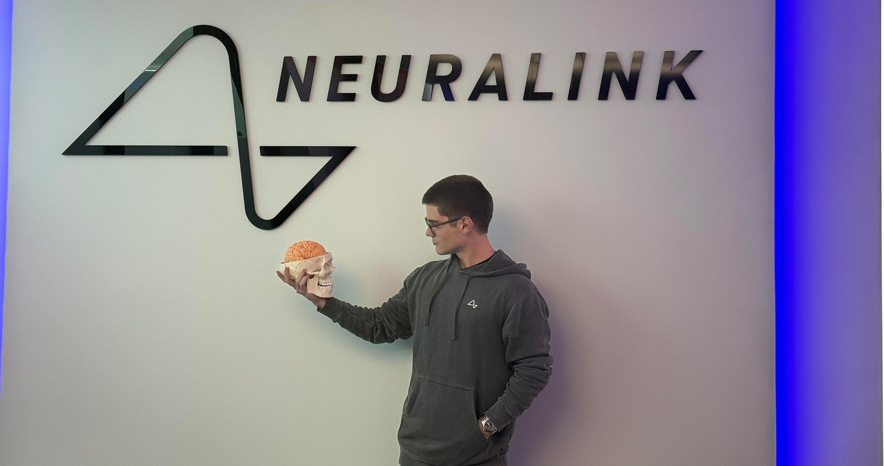 Matthew Mateo stands in front of the Neuralink sign holding a fake skull with part of the brain exposed.